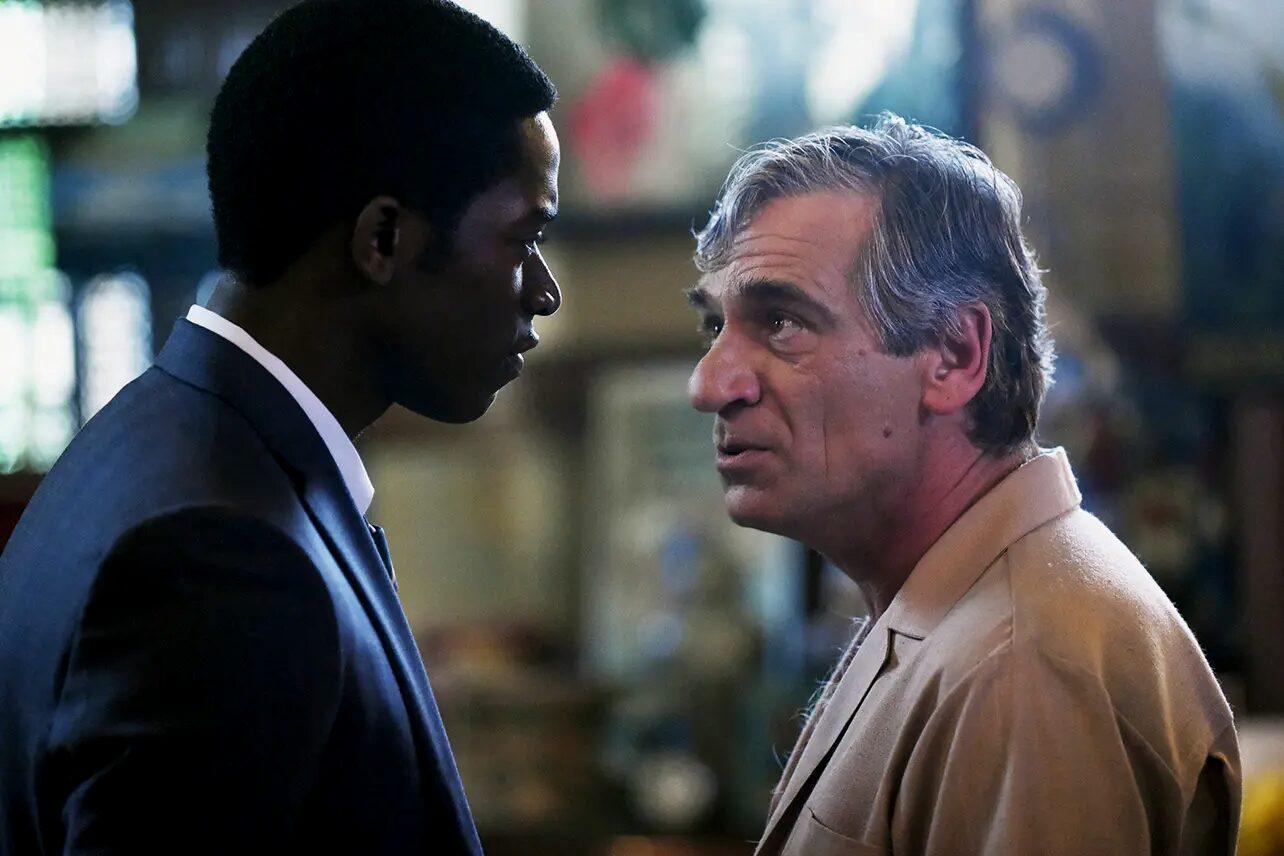 Two men in profile facing each other in close conversation: a young man in a dark suit and an older man in a tan jacket.