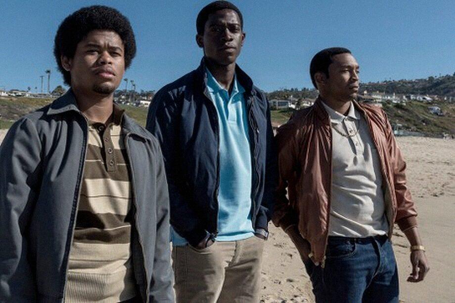 Three young men standing together on a Southern California beach, hills and houses behind them.