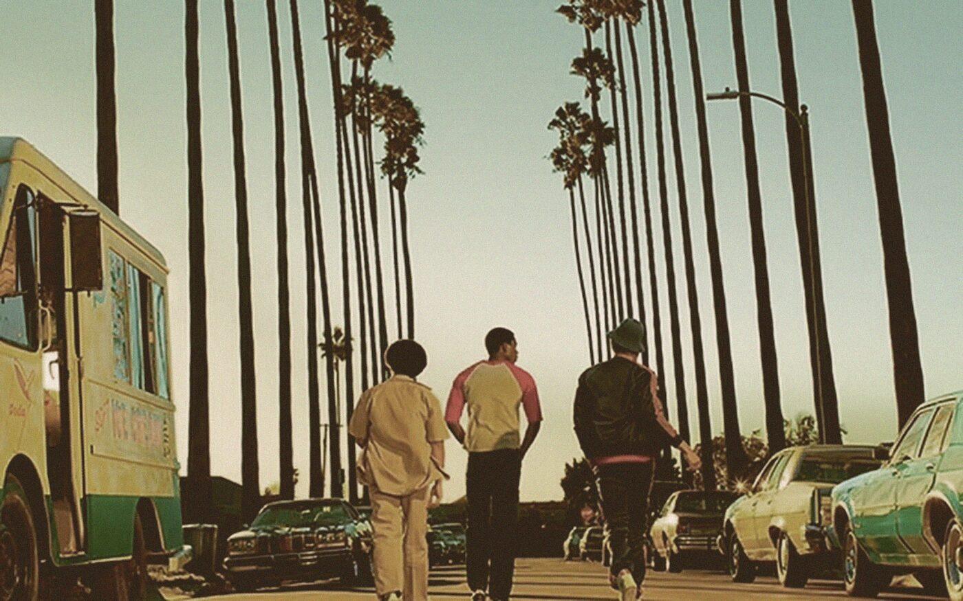 Three young men walking away down the middle of a palm-lined South Los Angeles street at golden hour, an ice cream truck and parked cars along the curb.