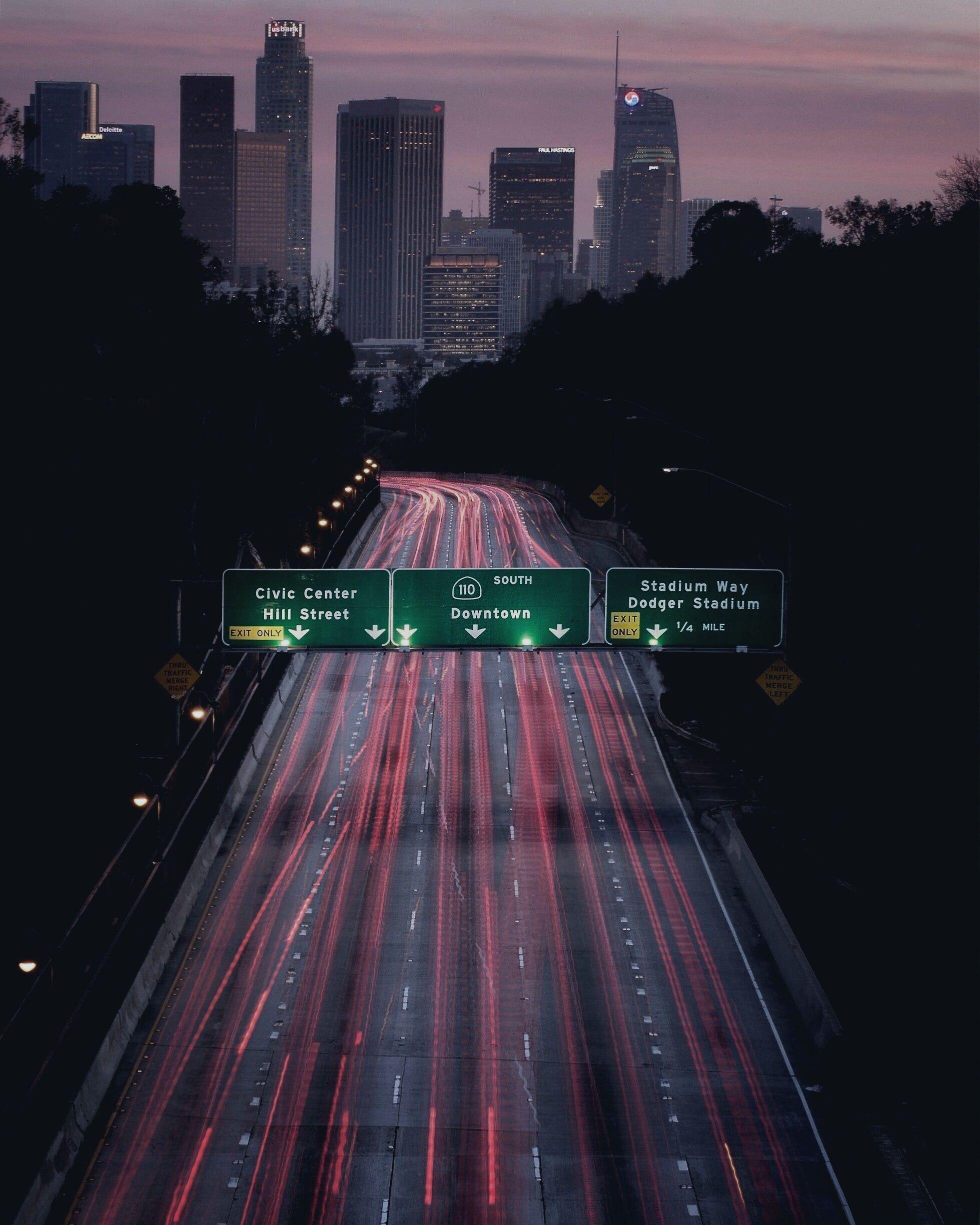 The 110 freeway running toward the downtown Los Angeles skyline at dusk, headlights and taillights drawn into long streaks.