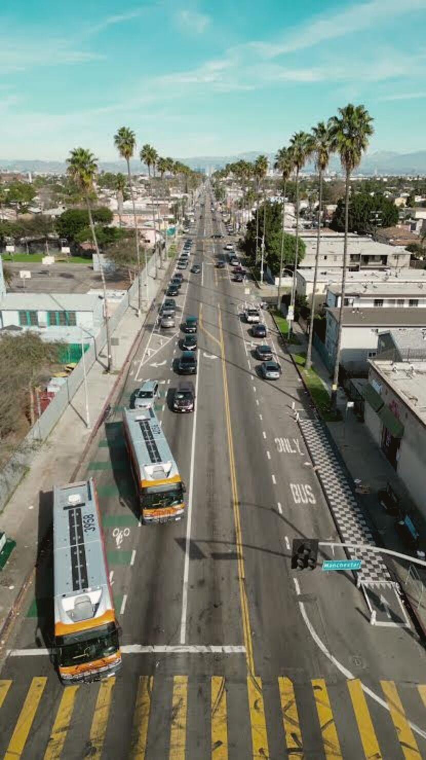 A Los Angeles boulevard from above at midday: two city buses, traffic, low storefronts, palms and mountains beyond.